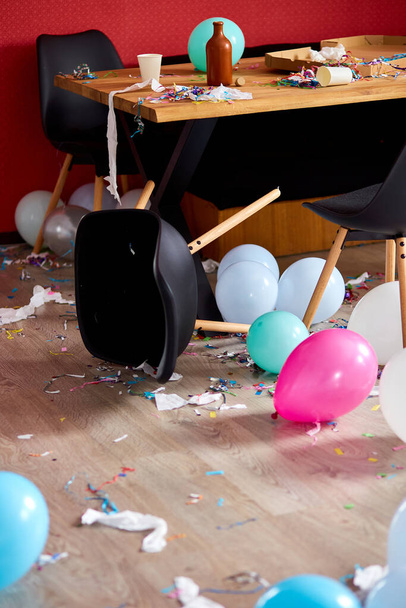 Après le chaos de la fête, désordonné dans le salon à la maison, table avec des pizzas et des verres à champagne couverts de confettis et de ballons, chaise sur le sol le matin après la fête. - Photo, image