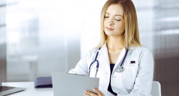 Young optimistic woman-doctor is using a clipboard, while sitting at the desk in her cabinet in a clinic. Portrait of friendly female physician woman with a stethoscope. Perfect medical service in a - Foto, imagen