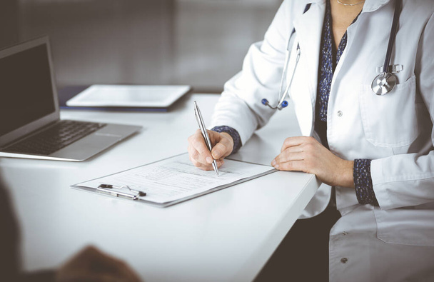 Unknown woman-doctor is writing some medical recommendations to her patient, while they are sitting together at the desk in the cabinet in a clinic. Physician is using a clipboard, close-up. Perfect - Photo, image