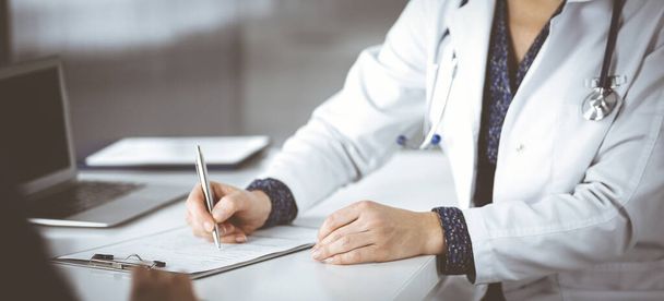 Unknown woman-doctor is writing some medical recommendations to her patient, while they are sitting together at the desk in the cabinet in a clinic. Physician is using a clipboard, close-up. Perfect - Foto, afbeelding