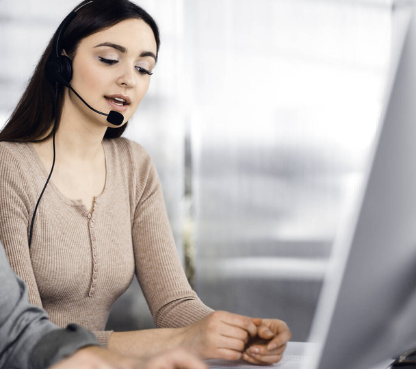Two young people in headsets are talking to the clients, while sitting at the desk in an office. Focus on woman. Call center operators at work - Photo, image