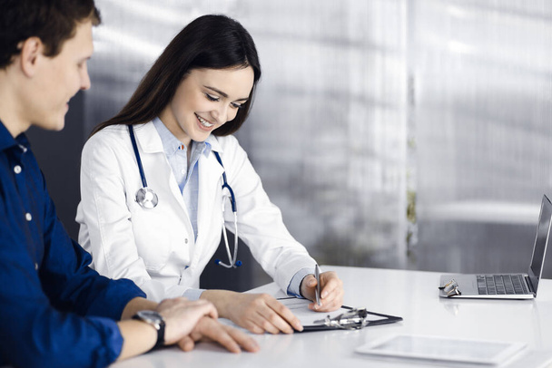Friendly young woman-doctor is listening to her patient, while they are sitting together at the desk in a cabinet. Physician is holding a clipboard in her hands for filling up medication - Foto, Imagen