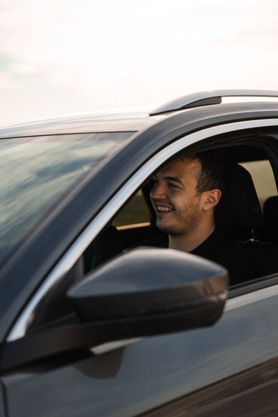 Homme assis sur le siège d'une nouvelle voiture, souriant en regardant vers l'avant, portant des vêtements sombres. Homme caucasien aux cheveux foncés et à la barbe courte. Dans la soirée dans une voiture gris foncé. - Photo, image
