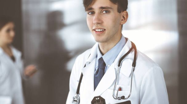 Friendly young male doctor standing with clipboard in modern clinic. Female colleague is busy with filling up of medication records form at the background of physician. Medicine concept - Foto, afbeelding