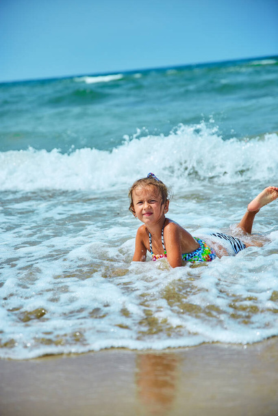 Счастливая маленькая девочка лежит на берегу моря в волнах во время летнего отдыха на пляже, ребенок, детская игра на море, летнее время, отдых resot. - Фото, изображение