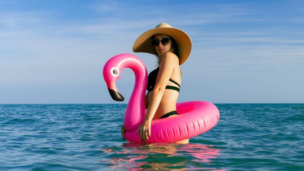 リゾートウェルネス。海のビーチの背景に青い海の水にピンクのインフレータブルフラミンゴとビキニ水着、サングラスやわらの帽子で幸せな若いセクシーな女の子。リゾート休暇で - 写真・画像