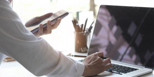 Крупним планом молода жінка використовує мобільний телефон для пошуку інформації під час роботи з ноутбуком на столі
 - Фото, зображення