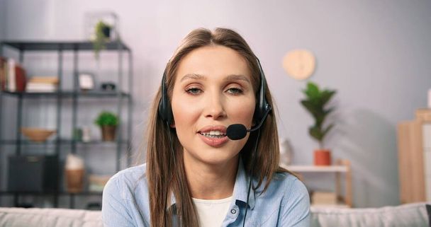 Portrait view of the focused young caucasian woman in headset with microphone sitting on sofa, involved in video call conversation. Long haired girl having online meeting workshop with colleagues - Photo, image