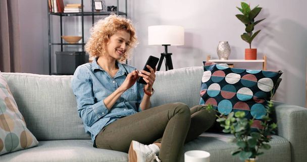 Smiling curly girl relaxe while sitting on sofa at home and using her modern cellphone. Blonde girl chatting with friends. Happy young woman on couch holding smartphone while check mobile application - Zdjęcie, obraz