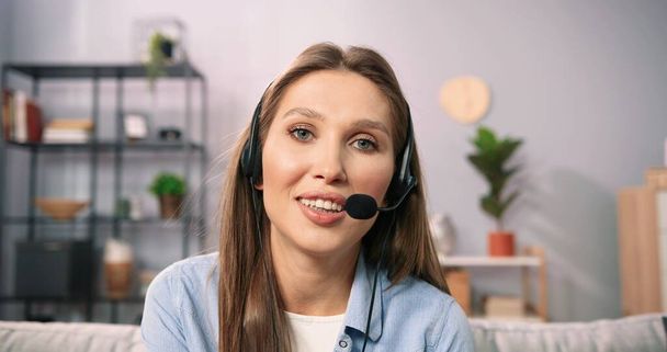 Portrait view of the focused young caucasian woman in headset with microphone sitting on sofa, involved in video call conversation. Long haired girl having online meeting workshop with colleagues - Foto, imagen