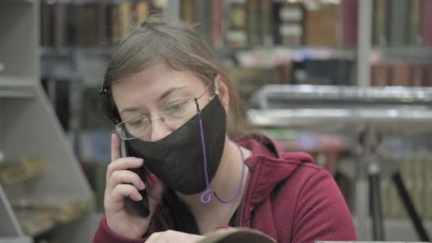 Γυναίκα με μάσκα και γυαλιά γυρίζει μέσα από το βιβλίο στη βιβλιοθήκη και μιλάει στο τηλέφωνο - Πλάνα, βίντεο