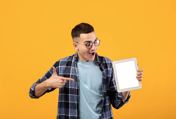 Turuncu stüdyo arka planında beyaz ekranlı tablet gösteren milenyum adamı şok etti, web sitesi veya uygulama için alan - Fotoğraf, Görsel