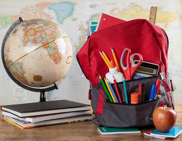 Book bag with an assortment of school supplies on a desk with world map in background - Photo, Image