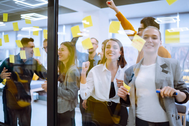 Group of young modern people working on project together. Business colleagues writing on sticky note on glass wall with coworkers standing by in office. Startup Business. Planning, analysis. - Foto, imagen