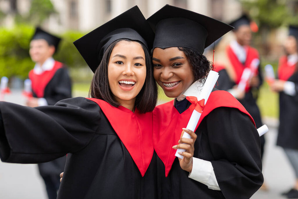 Joyeux jeunes femmes en robes de remise des diplômes prenant selfie - Photo, image