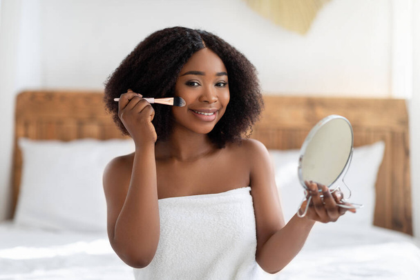 Mulher afro-americana atraente olhando no espelho, aplicando maquiagem na cama em casa. Conceito de rotina diária - Foto, Imagem