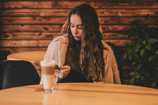 Μια γυναίκα με μοντέρνα εμφάνιση με μακριά μαλλιά κάθεται σε ένα τραπέζι σε ένα εστιατόριο με ένα τηλέφωνο στα χέρια της - Φωτογραφία, εικόνα