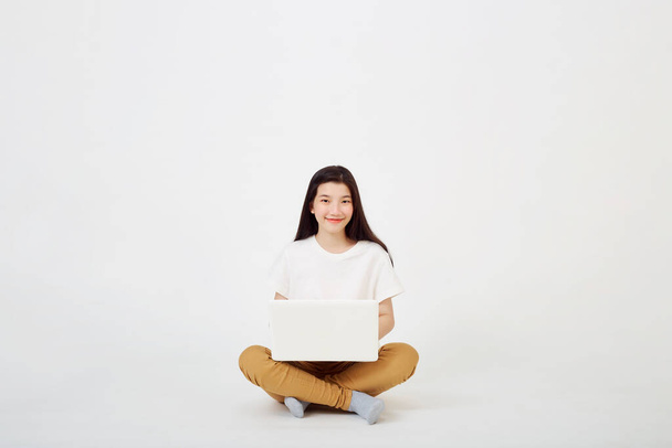 Счастливая молодая азиатская женщина красиво сидит на полу со скрещенными ногами и использует ноутбук на заднем плане - Фото, изображение