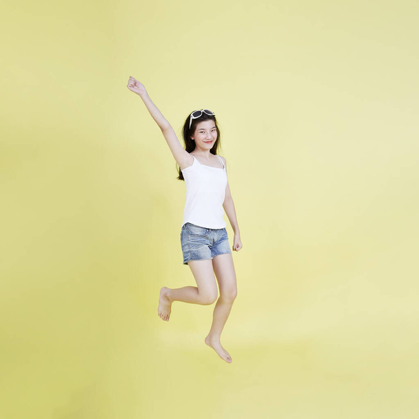 Volledig portret van mooi Aziatisch meisje zwevend lachend en springend in de lucht met de hand omhoog gericht geïsoleerd op heldere gele achtergrond, met kopieerruimte - Foto, afbeelding