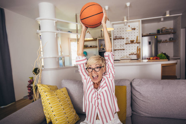 Ηλικιωμένη γυναίκα συναισθηματικά βλέποντας μπάσκετ στην τηλεόραση και γιορτάζοντας τη νίκη στο σπίτι. - Φωτογραφία, εικόνα