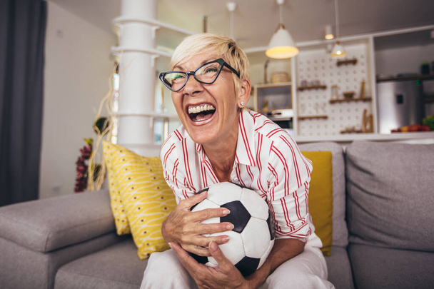 Starsza kobieta emocjonalnie ogląda piłkę nożną w telewizji i świętuje zwycięstwo w domu. - Zdjęcie, obraz