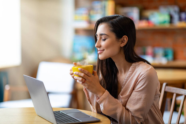 Задоволена арабська фрілансерка насолоджується ранковою кавою в кафе, працює онлайн на ноутбуці
 - Фото, зображення