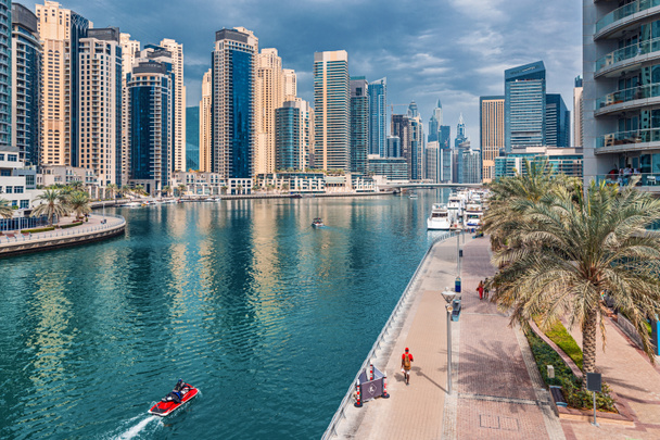 Moderní a rozvinutá oblast Dubai Marina s vysokými mrakodrapy a komerčními a rezidenčními nemovitostmi. Na vodě se vznáší vodní skútr a lidé kráčejí podél náspu - Fotografie, Obrázek