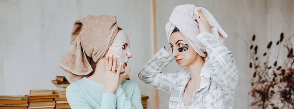 Two girls make masks and patches for the beauty of face and hair. Women take care of youthful skin. Girlfriends laugh, make faces and do beauty treatments at home with towels on their heads. - Foto, afbeelding