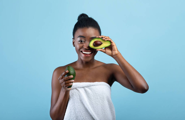Beautiful african american lady wrapped in towel covering eye with half of avocado over blue studio background - Фото, изображение