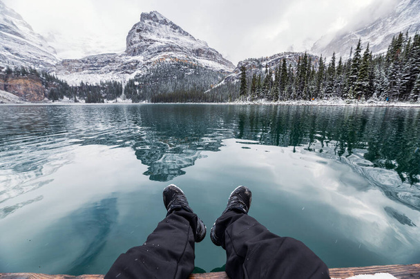 Nogi podróżnika relaks na molo z odbiciem skalistych gór w jeziorze O 'Hara w zimie w Parku Narodowym Yoho, Kanada - Zdjęcie, obraz