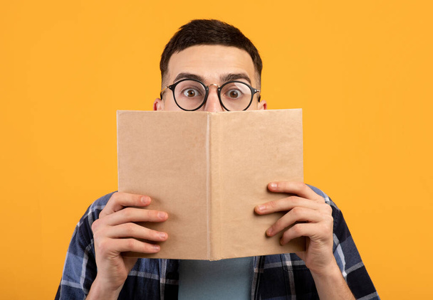Shocked young guy in glasses hiding behind book, having problem studying for college exams on orange background - Фото, изображение