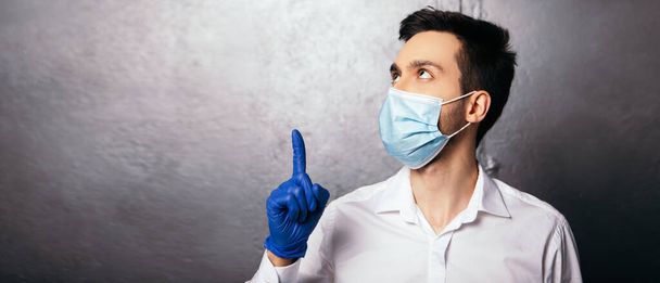 L'homme montre l'index vers le haut geste. Médecin infirmier vêtu d'une chemise blanche avec masque viral bleu et gants. Espace de copie - Photo, image