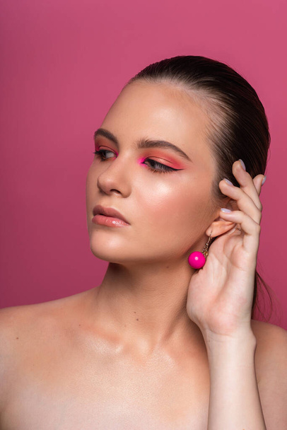 High beauty photo of a lovely young girl with wonderful professional makeup, round pink earrings, with long brown hair. Posing over rosy background. Close-up. Studio shot. Portrait - Fotografie, Obrázek