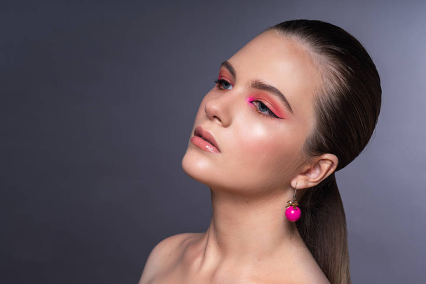 High beauty photo of a lovely young girl with wonderful professional makeup, round pink earrings, with long brown hair. Posing over grey background. Close-up. Studio shot. Portrait - Фото, изображение