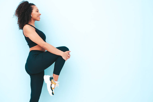 Portrait of fitness smiling black woman in sports clothing with afro curls hairstyle.She wearing sportswear.Young beautiful model stretching out before training.Female sitting in studio near blue wall - Foto, immagini