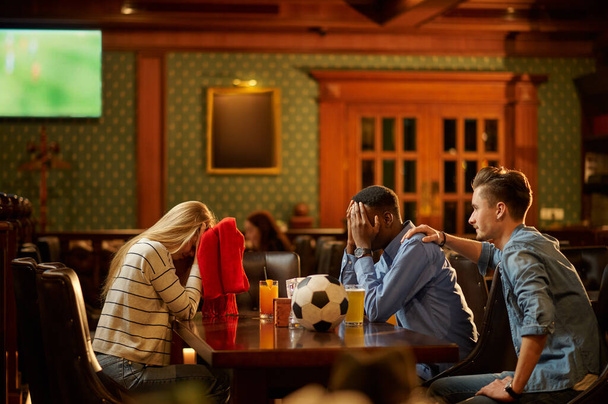 Fans de football avec écharpe rouge regarder la traduction du jeu, amis dans le bar. Groupe de personnes se détendre dans le pub, mode de vie nocturne, amitié, célébration sportive - Photo, image