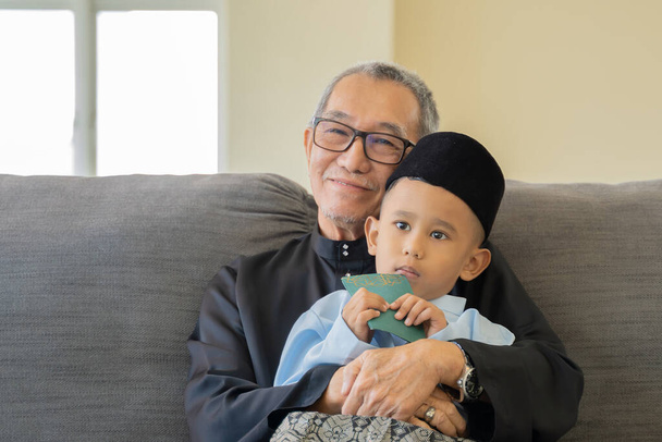 Momento de celebración de Eid Mubarak, abuelo con nieto vestido con tela tradicional en el sofá. - Foto, Imagen