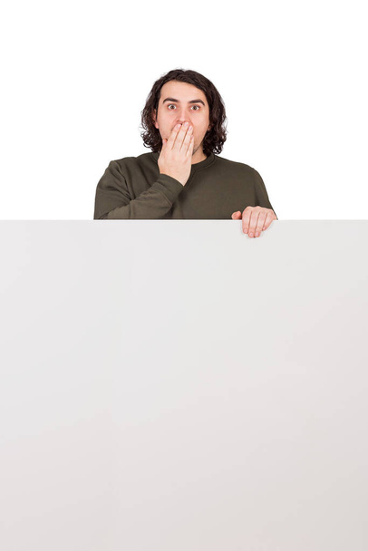 Shocked man, staring to camera with big eyes, covers his mouth with hand, stands behind a blank banner for announcements. Empty sheet with copy space for advertising and messages and astonished guy - Photo, Image