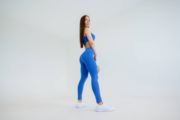 Sexy young girl performs exercises with a weight in a blue tracksuit on a white background. Fitness, healthy lifestyle - Fotoğraf, Görsel