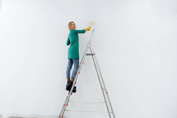 Merdivendeki genç bir kız beyaz bir duvarı silindirle boyuyor. İç kısmın tamiri. - Fotoğraf, Görsel
