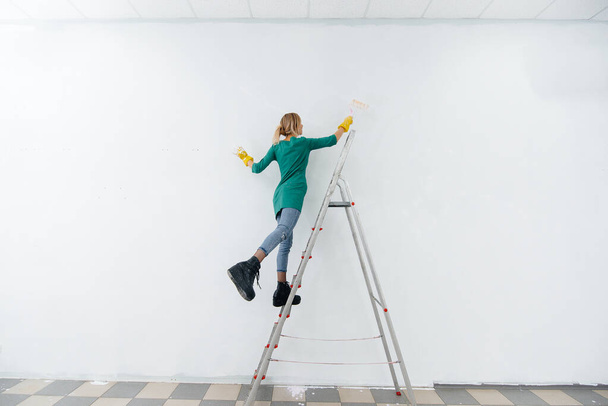 A young girl on a stepladder paints a white wall with a roller. Repair of the interior. - Photo, Image