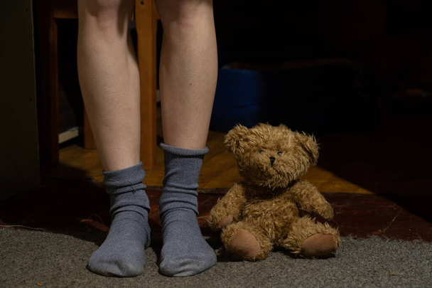 γυναικεία πόδια σε κάλτσες και καφέ αρκουδάκι στο πάτωμα σε σκοτεινό δωμάτιο - Φωτογραφία, εικόνα
