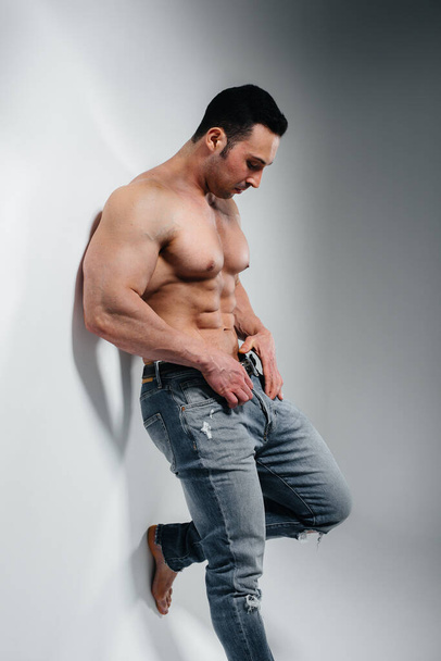 A young athlete bodybuilder poses in the studio topless in jeans near the wall. Sport. - Photo, Image