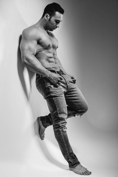 A young athlete bodybuilder poses in the studio topless in jeans near the wall. Black and white. - 写真・画像