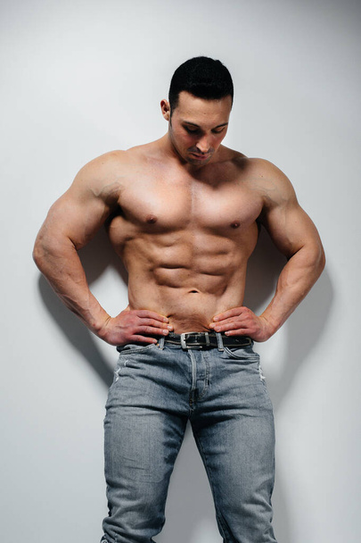 A young athlete bodybuilder poses in the studio topless in jeans near the wall. Sport. - Фото, зображення