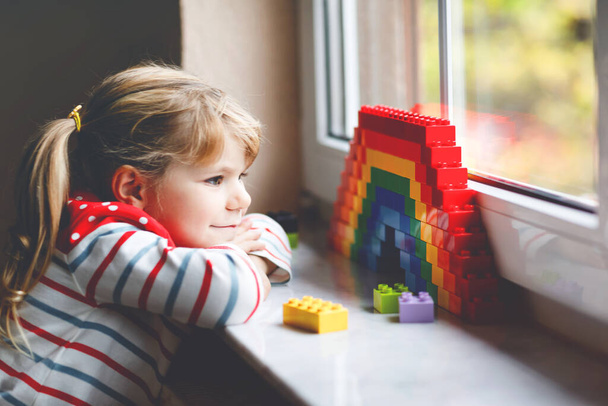 Милая маленькая девочка за окном создает радугу с красочными пластиковыми блоками во время пандемического карантина коронавируса. Дети делают и рисуют радуги по всему миру в знак. - Фото, изображение