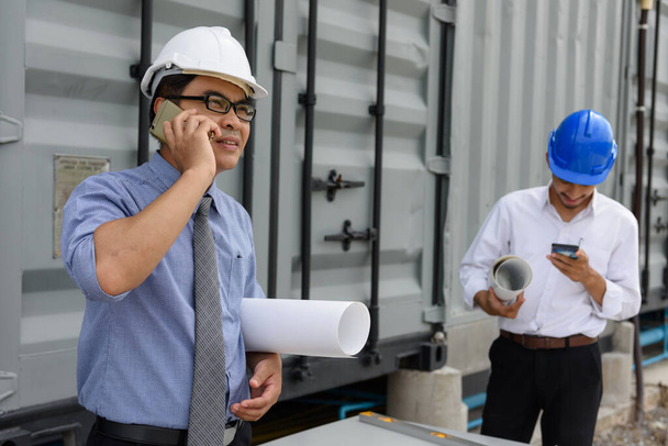 Asian Engineer senior manager hold blueprint and talk over phone at construction site while young engineer reading project plan. Engineering communication by 4g technology. - Photo, Image