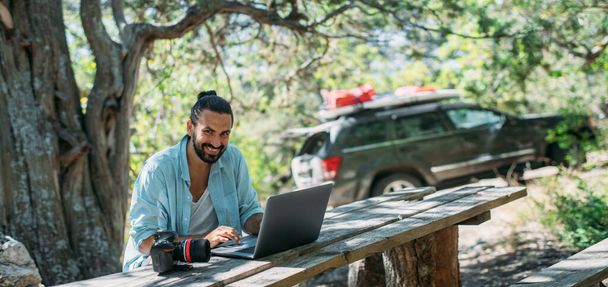 Ein männlicher Fotograf arbeitet auf einem Laptop im Freien auf einem Campingplatz. Junger Mann, Freiberufler, Reiseblogger, der online am Laptop arbeitet, während er im Freien in den Bergen wandert. Arbeit von überall auf der Welt. - Foto, Bild