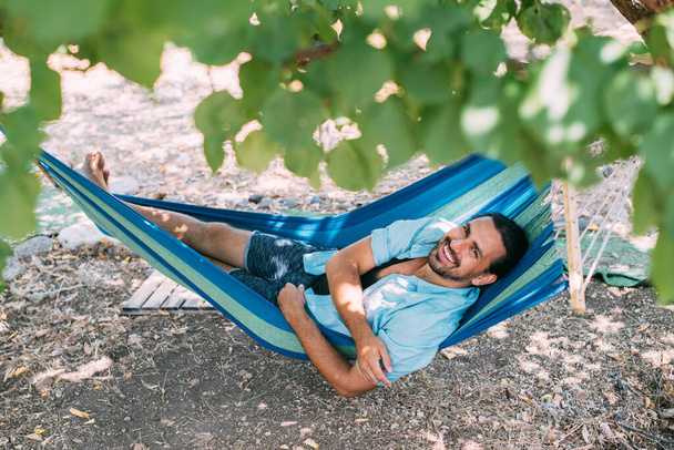 Nuori mies lepää riippumatossa puutarhassa. Komea kaveri makaa rentona riippumatossa puutarhapuiden varjossa kesäpäivänä. - Valokuva, kuva