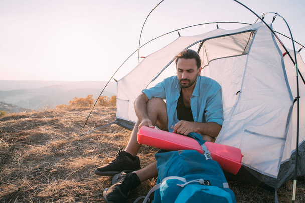 Young male tourist with a backpack in a tent in the mountains. A guy breaks camp on a hike, sits under an awning with a large backpack and sleeping foam alone in the mountains at sunset - Photo, Image
